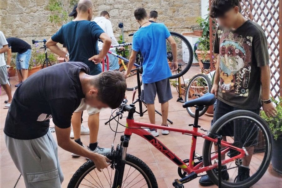 Dos jóvenes arreglan la rueda de una bicicleta mientras al fondo otros jóvenes siguen las indicaciones de los monitores